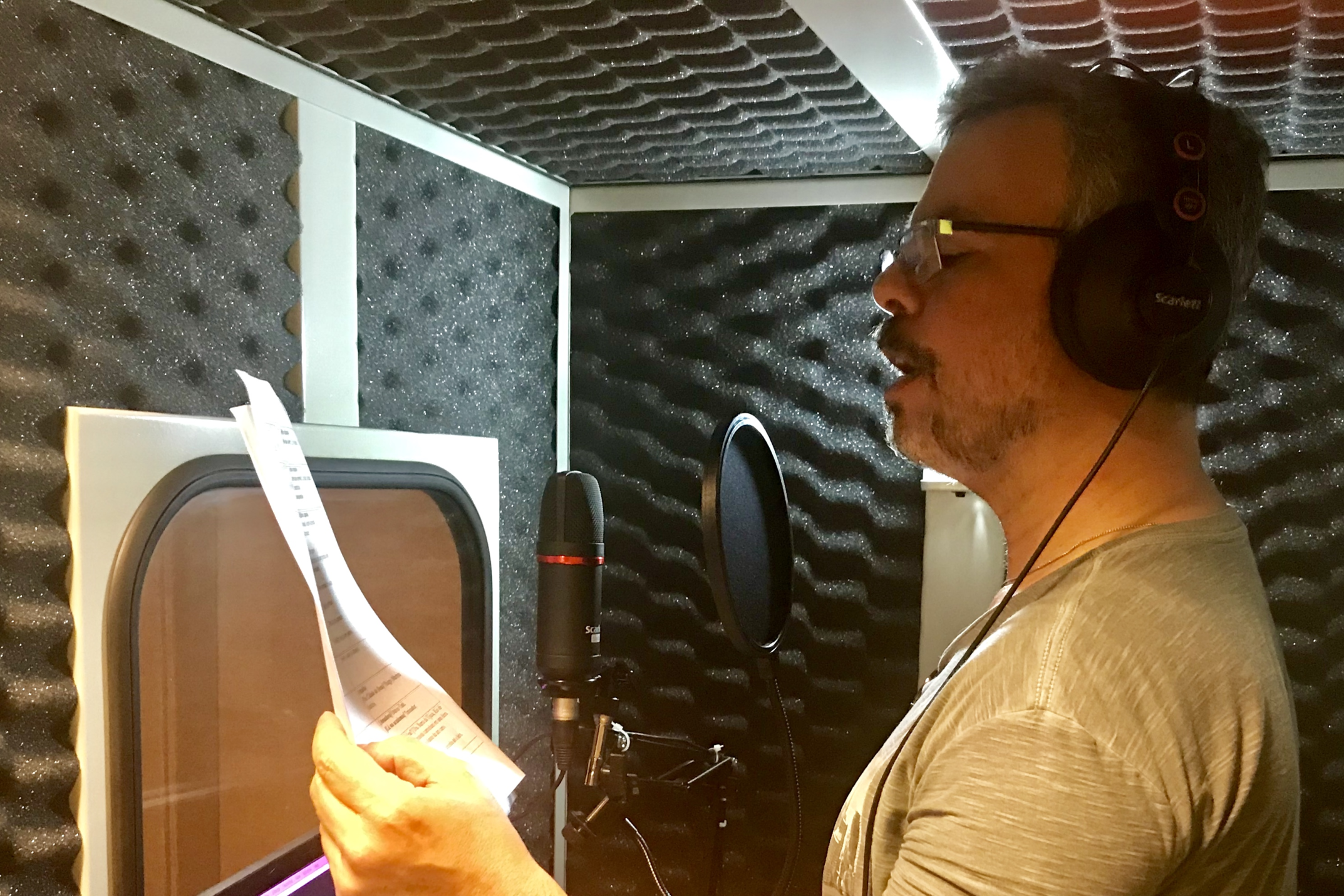 Homem de óculos e fones de ouvido cinza gravando narração de audiodescrição em um estúdio com isolamento acústico em formato de caixa de ovo, segurando um roteiro impresso em frente a um microfone profissional.
