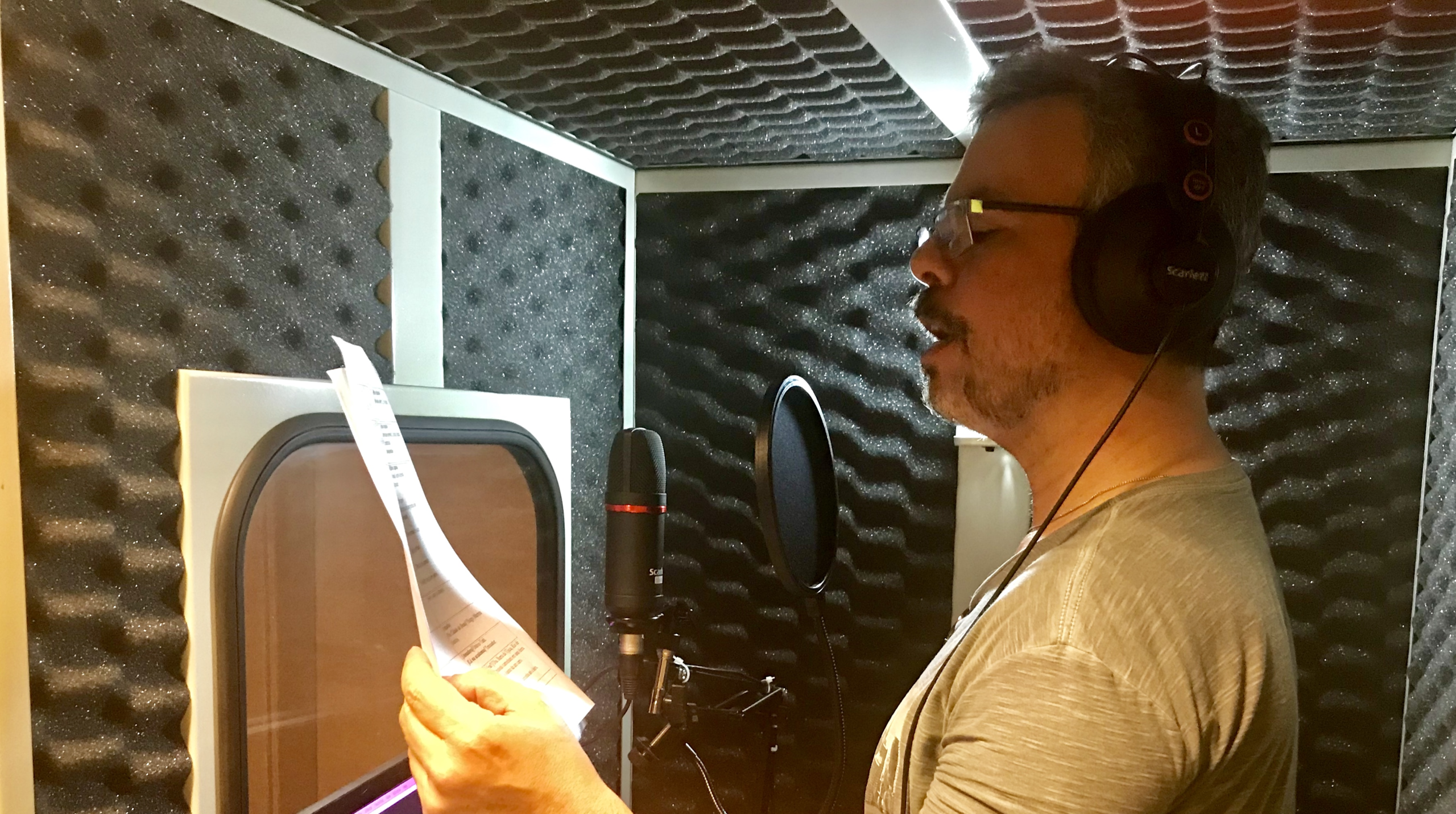 Homem de óculos e fones de ouvido cinza gravando narração de audiodescrição em um estúdio com isolamento acústico em formato de caixa de ovo, segurando um roteiro impresso em frente a um microfone profissional.